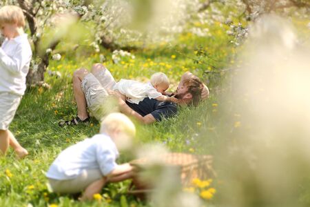A happy young father is laying in the flowers relaxing with his baby daughter as his two boy children play under the apple orchard trees after a picnic on a spring day.の写真素材