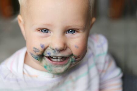 a happy, laughing one year old baby girl has a messy face covered in blue and purple paint.の写真素材