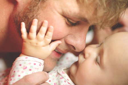 A happy father is playing with his newborn baby daughter, lovingly touching noses while she puts her...
