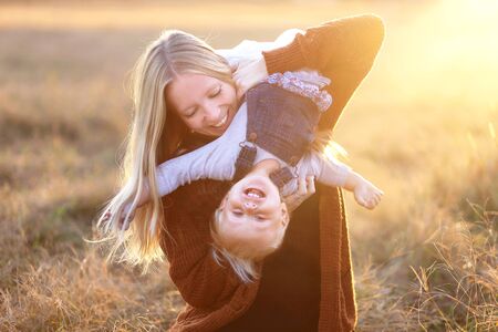 A happy young mother is playing outside with her one and a half year old baby daughter, holding her upsidedown and laughing at sunset.の写真素材