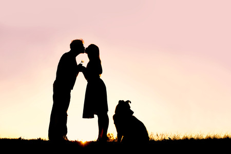 A silhouette of a happy young married couple in love, kissing at sunset, next  to their pet dog outside on a summer night.の写真素材