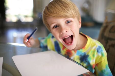 A super happy and excited little boy with a big smile is looking at the camera as he draws with pen on a white paper.の写真素材