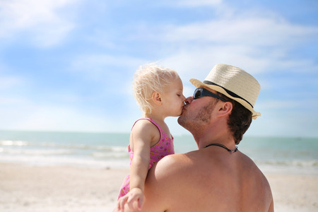 A young father is lovingly kissing his one year old baby daughter as they relax on the beach by the ocean.の写真素材