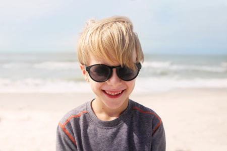 A happy, smiling little 7 year old boy is laughing as he stands on the beach by the ocean on a windy summer day.の写真素材