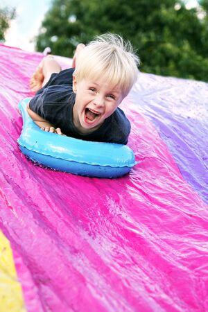 A happy young child is screaming as he slides down a slip-and-slide in his backyard.の写真素材
