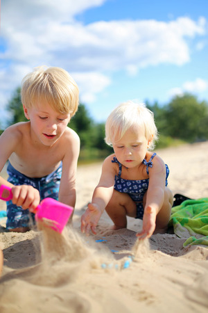 Two happy little children, a boy and his baby sister, are playing in the sand at the beach, building a sandcastle.の写真素材