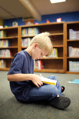 A little second grade elementary school aged boy child is sitting on the carpet, reading a book at the school library.の写真素材