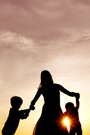 A silhouette of a happy mother and her two little boy children holding hands and dancing outside at sunset.の写真素材