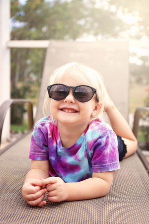 A cute, 2 year old toddler child is smiling as she poses for the camera on a beach chair under the creen by the pool on a summer day.の写真素材