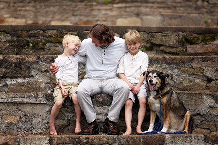 A young, loving father is sitting outside on stone steps, with his two boy children and their adopted pet German Shepherd dog on a Summer day.の写真素材