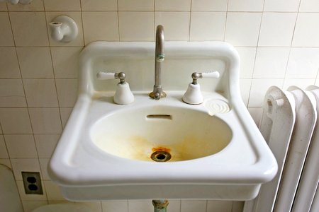 A white tile wall background behind an old vintage, original 1920's farmhouse porcelain bathroom sink with rust stains on the drain.の写真素材