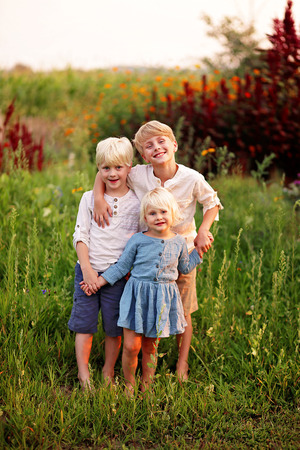 Three sweet blond farm children are smiling as they pose for a family portrait in a country garden.の写真素材