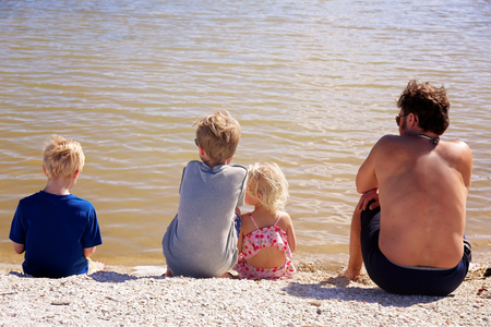 A family of a father and his three young children are sitting together on the beach by the ocean looking out at the water on a sunny summer day.の写真素材