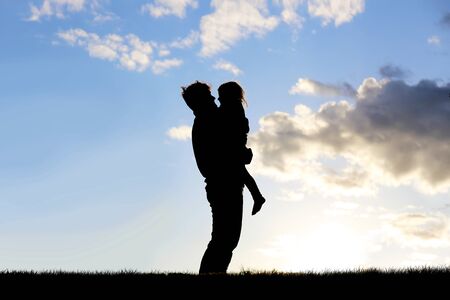 The silhouette of a happy young father holding his little girl child and hugging her as they stand outside at dusk.の写真素材