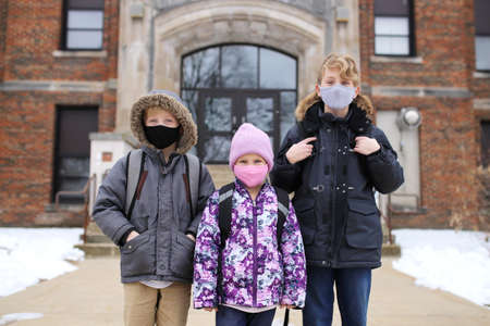 Three little kids are standin in front of their elemtary school building, wearing backpacts and...