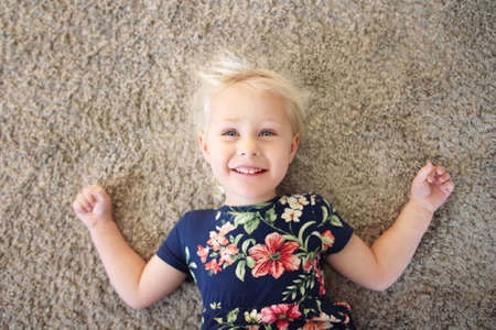 A happy little girl child in a flower dress is laying on her home carpet smiling.