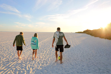 Three children, siblings, are walking on a large white sand beach, toward a beautiful sunset over...