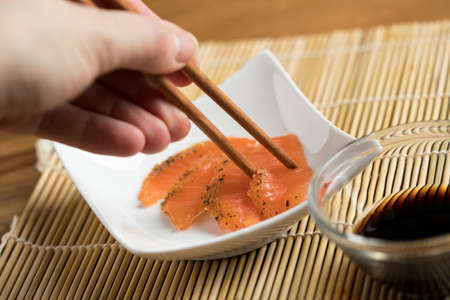 Woman eating smoked and stained salmon sashimi slices on porcelain plate with soy sauce using chopsticksの写真素材