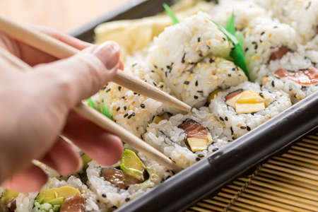 Woman eating salmon, avocado and mango inside out California sushi with wooden chopsticks from take away Bento boxの写真素材