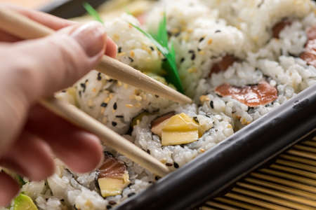 Woman eating salmon, avocado and mango inside out California sushi with wooden chopsticks from take away Bento boxの写真素材