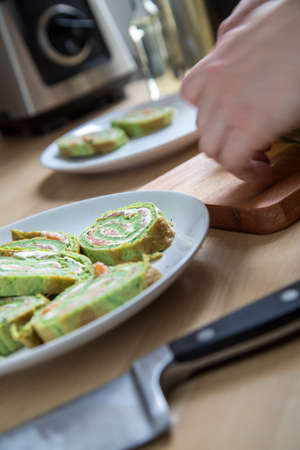 Woman cut and arrange spinach pancake roll with smoked salmon and cream cheese in domestic kitchenの写真素材