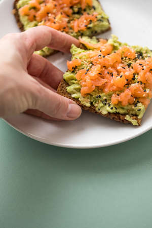 Woman eating avocado smoked salmon on sunflower seed whole grain bread slice with sesame seeds for breakfast or snack on mint green backgroundの写真素材