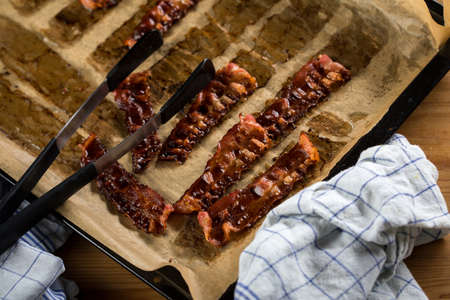 Crispy fried bacon rashers on oven tray and baking paper with towel and pliers in domestic kitchen