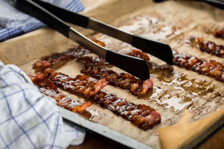 Crispy fried bacon rashers on oven tray and baking paper with towel and pliers in domestic kitchenの写真素材
