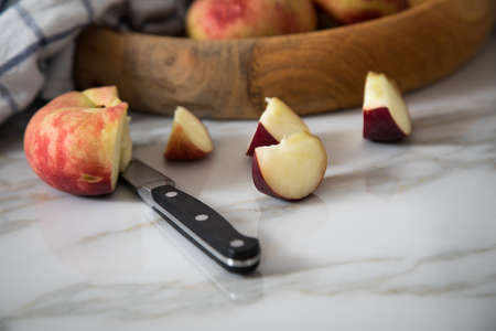 Flat peaches with knife and towel in wooden bowl on marble table
