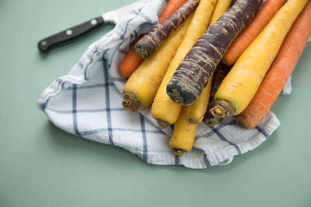Colorful rainbow carrots in purple, orange and yellow on kitchen towel with knife on pastel mint green backgroundの写真素材