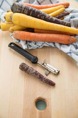 Colorful rainbow carrots in purple, orange and yellow on kitchen towel with peeler on wooden boardの写真素材