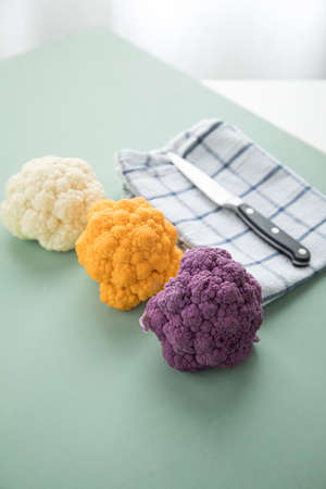 Colorful rainbow cauliflower in purple, orange and white on kitchen towel with knife on pastel green...