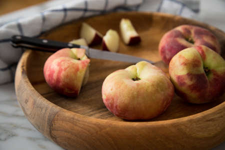 Flat peaches with knife and towel in wooden bowl on marble tableの写真素材