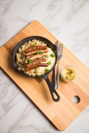 Small fried Bavarian German NÃ¼rnberger sausages with sauerkraut, mashed potatoes and mustard in iron cast pan on marble tableの写真素材