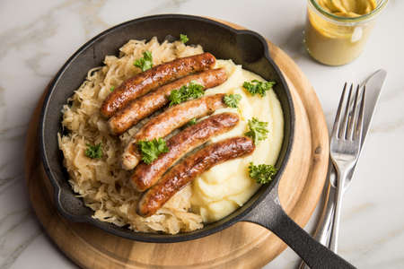 Small fried Bavarian German NÃ¼rnberger sausages with sauerkraut, mashed potatoes and mustard in iron cast pan on marble tableの写真素材