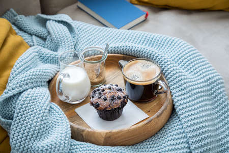 Blueberry muffin with coffee, sugar and milk on wooden tray and comfy sofa with blue wool knit blanket, yellow cushions and bookの写真素材