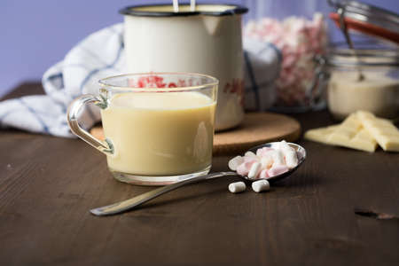 Preparation and cooking of hot white chocolate in milk pot with instant powder and marshmallows in domestic kitchen on dark wooden counter top and lilac background