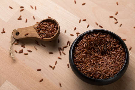 Red skinned raw Jasmine rice in black bowl with spoon on wooden background and is a specialty from Cambodia Asiaの写真素材