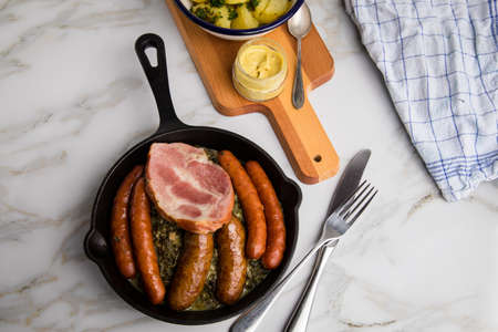 Traditional German Oldenburger green cabbage with smoked sausage mix, Kassler pork neck, mustard and boiled potatoes in cast iron pan on marble tableの写真素材
