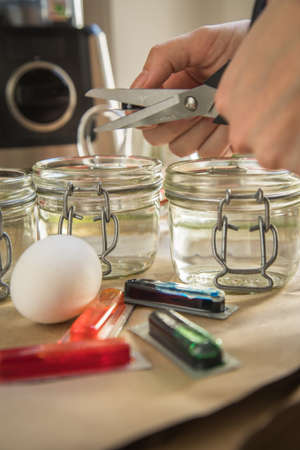 Woman pouring liquid red Easter cold color color in glass with water and vinegar in domestic kitchen next to orange, yellow, green, blue dye in packaging with boiled eggs and scissors for DIY coloringの写真素材