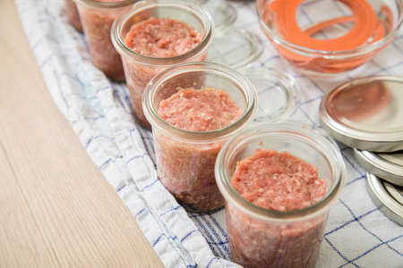 Raw mixed minced meat with pork and beef in screw-cap and preserving mason glass jars on towel for home-made pressure canning in domestic kitchen for long term food storage in pantryの写真素材