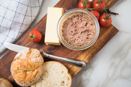 Canned liver sausage spread from long term pantry with crispy bread roll bun, butter, tomatoes and knife for snack or breakfast on wooden board and light marble backgroundの写真素材