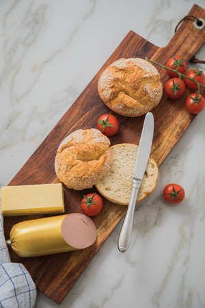 German fine veal liver sausage spread with two crispy bread rolls buns, tomatoes, butter and knife on wooden board, kitchen towel and light marble background for breakfastの写真素材