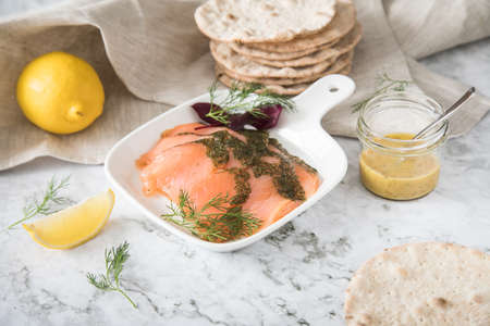 Gravlax a raw, marinated graved salmon with dill on plate with crispbread, Scandinavian mustard sauce, lemon and linen towel on light marble backgroundの写真素材