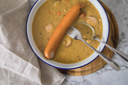 Hearty German yellow split pea soup stew with Bockwurst sausages in enamel dish on wooden board and light marble backgroundの写真素材