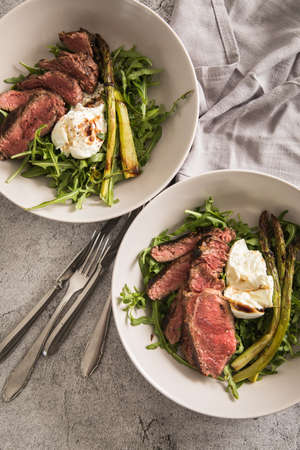 Roasted green asparagus with medium rare beef steak, rocked salad, buffalo mozzarella cheese and balsamic on baking tray, wooden board, cutlery and rustic dark stone backgroundの写真素材