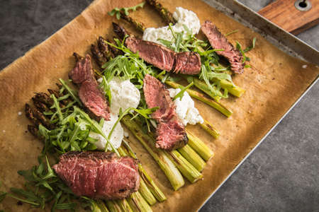 Roasted green asparagus with medium rare beef steak, rocked salad, buffalo mozzarella cheese and olive oil on baking tray, wooden board, cutlery and rustic dark stone backgroundの写真素材