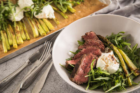 Roasted green asparagus with medium rare beef steak, rocked salad, buffalo mozzarella cheese and balsamic in bowl and baking tray, cutlery and rustic stone backgroundの写真素材