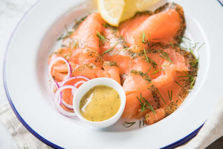 Gravlax a raw, marinated graved salmon with dill on enamel plate with Scandinavian mustard sauce, lemon, onion rings and linen towel on light marble backgroundの写真素材