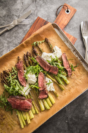 Roasted green asparagus with medium rare beef steak, rocked salad, buffalo mozzarella cheese and olive oil on baking tray, wooden board, cutlery and rustic dark stone backgroundの写真素材